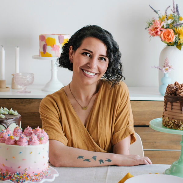 Whitney DePaoli sits at a table surrounded by four delicious cakes and several vases of flowers